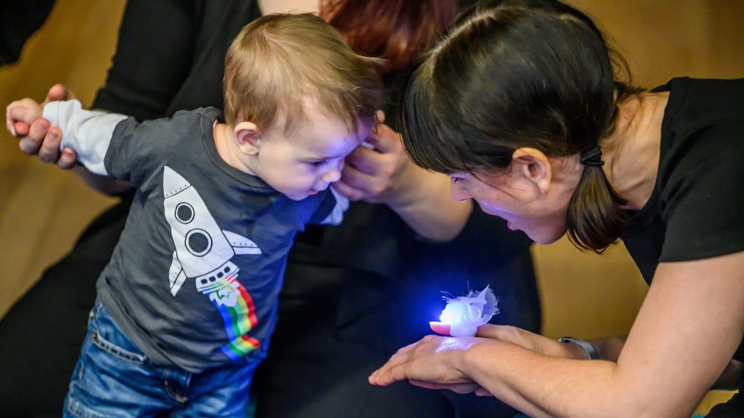 A toddler stares at a brightly lit shape on a performer's hand