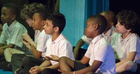 School boys in white shirts sat cross legged and watching something off to the left