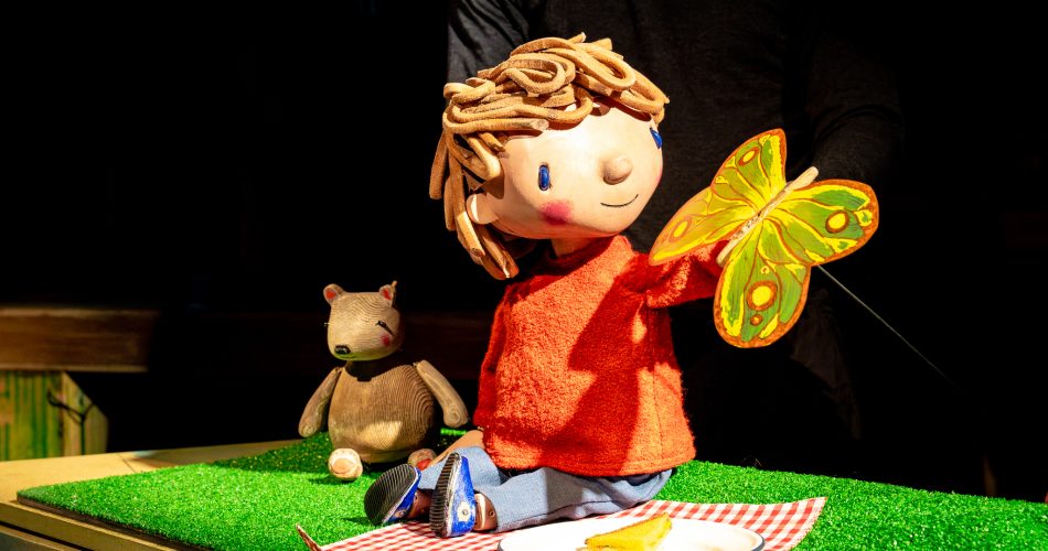 A puppet of a young boy in an orange jumper sits on a red and white checked picnic rug. A yellow butterfly lands on his hand.