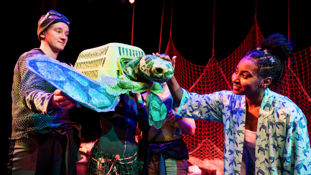 Two performers operate a puppet turtle made out of a washing basket while a third strokes it's head