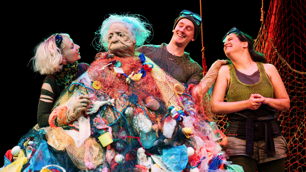 Three performers smile looking at a puppet elderly person covered in plastic waste