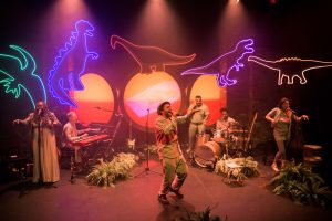 Five musicians play on stage. Behind them are various neon outlines of dinosaurs.