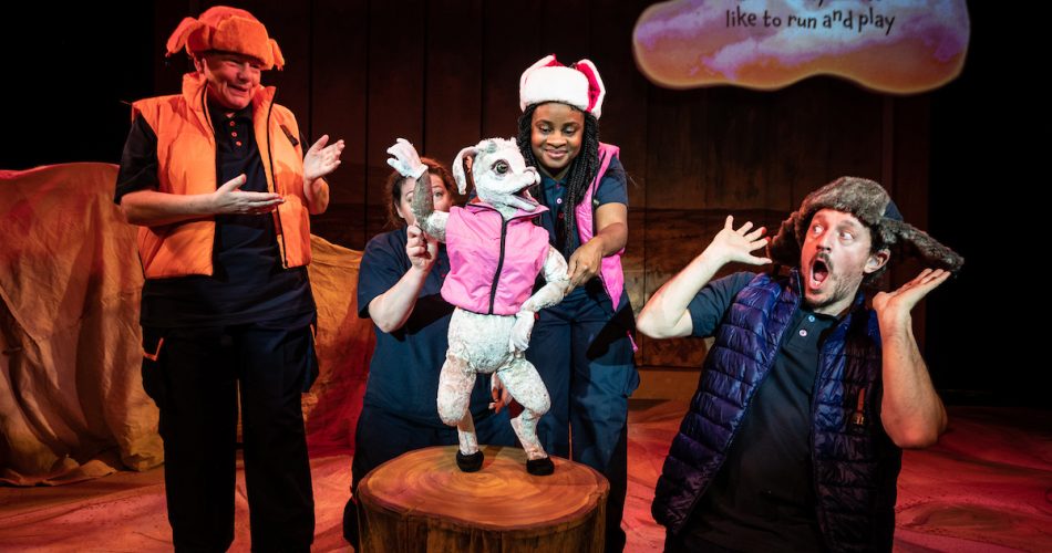 A puppeteer pilots a puppet of a goat in a pink gilet, a man looks on shocked