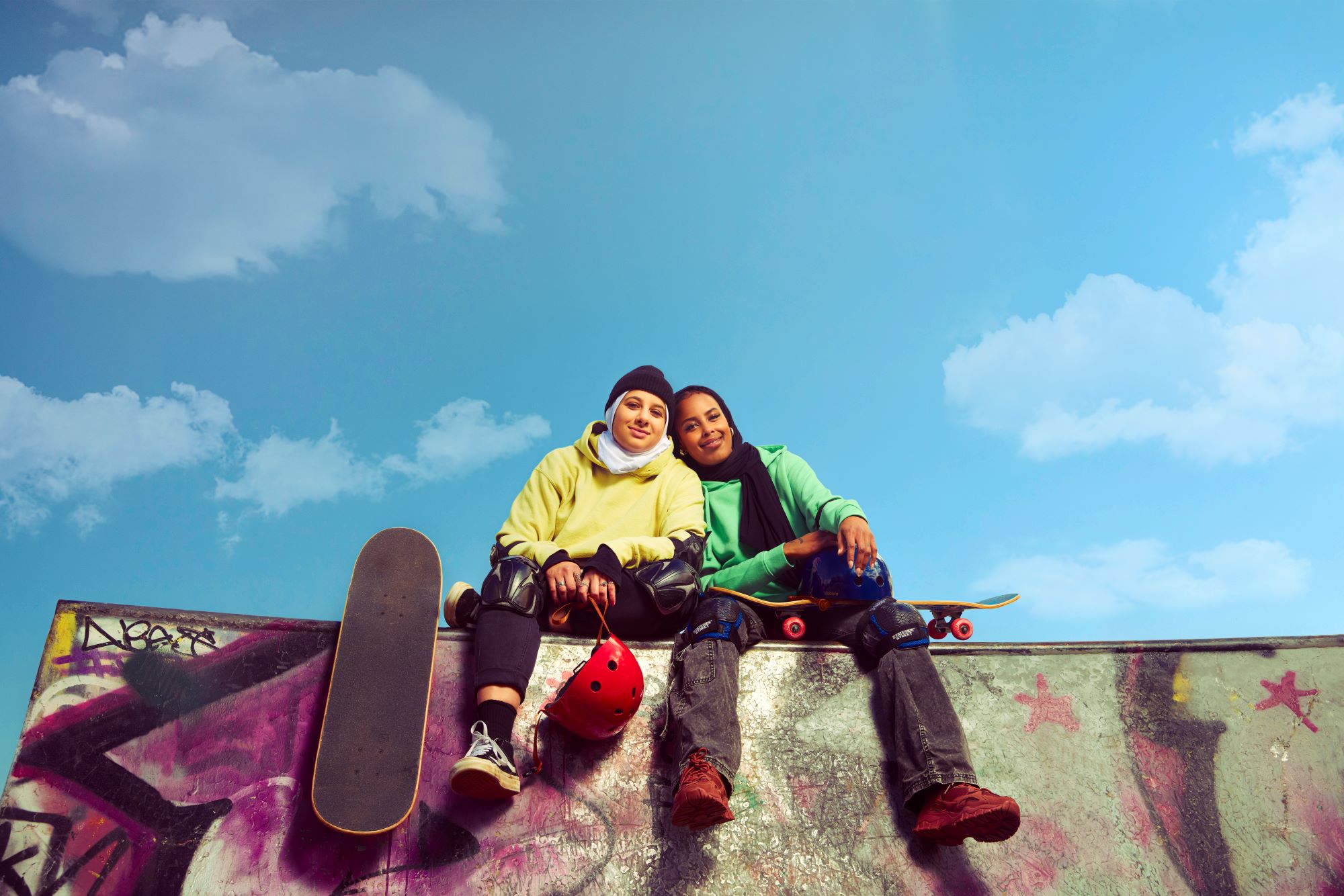 Two young women wearing hijabs, colourful hoodies and knee and elbow pads sit smiling together at the top of a skate ramp.