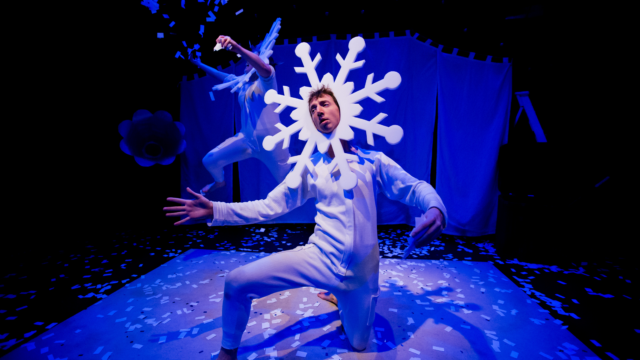 An adult performing on stage dressed in a white one-piece outfit wearing a snowflake headdress.