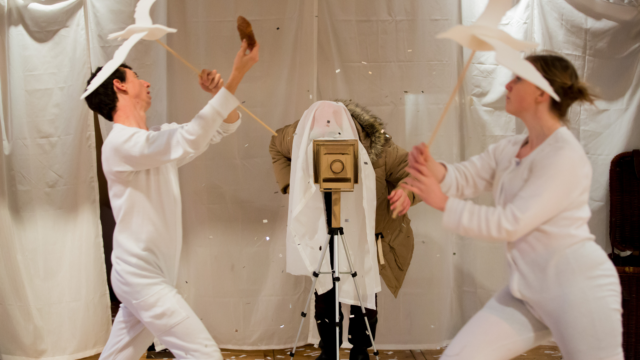 A couple of adults on stage dressed in white, holding white paper birds on wooden sticks.