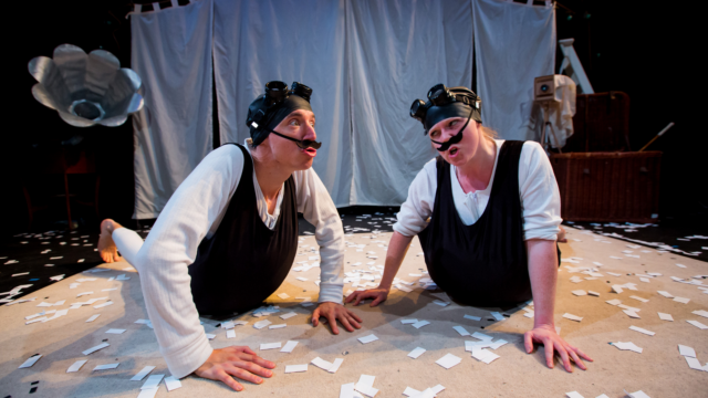 A couple of adults on stage with their hands touching the floor mid performance. They are wearing black goggles on their heads and fake black moustaches.