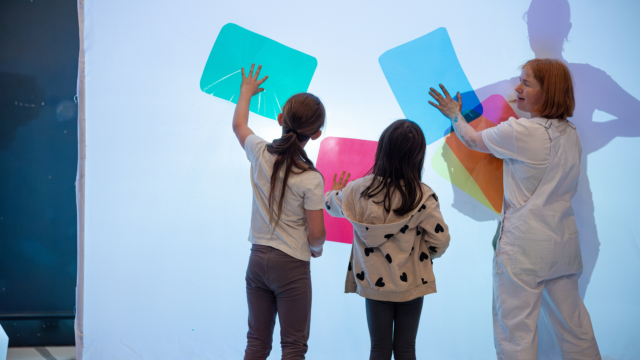 An adult wearing white dungarees with green paint splatters on them, stood in front of a projector with coloured shapes on. There are two children in front of the projecter with their hands on the shapes.