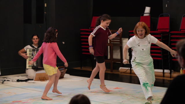 An adult wearing white dungarees with green paint splatters on them, being followed by two children who are running on a big sheet of white cloth that is splattered with colours.