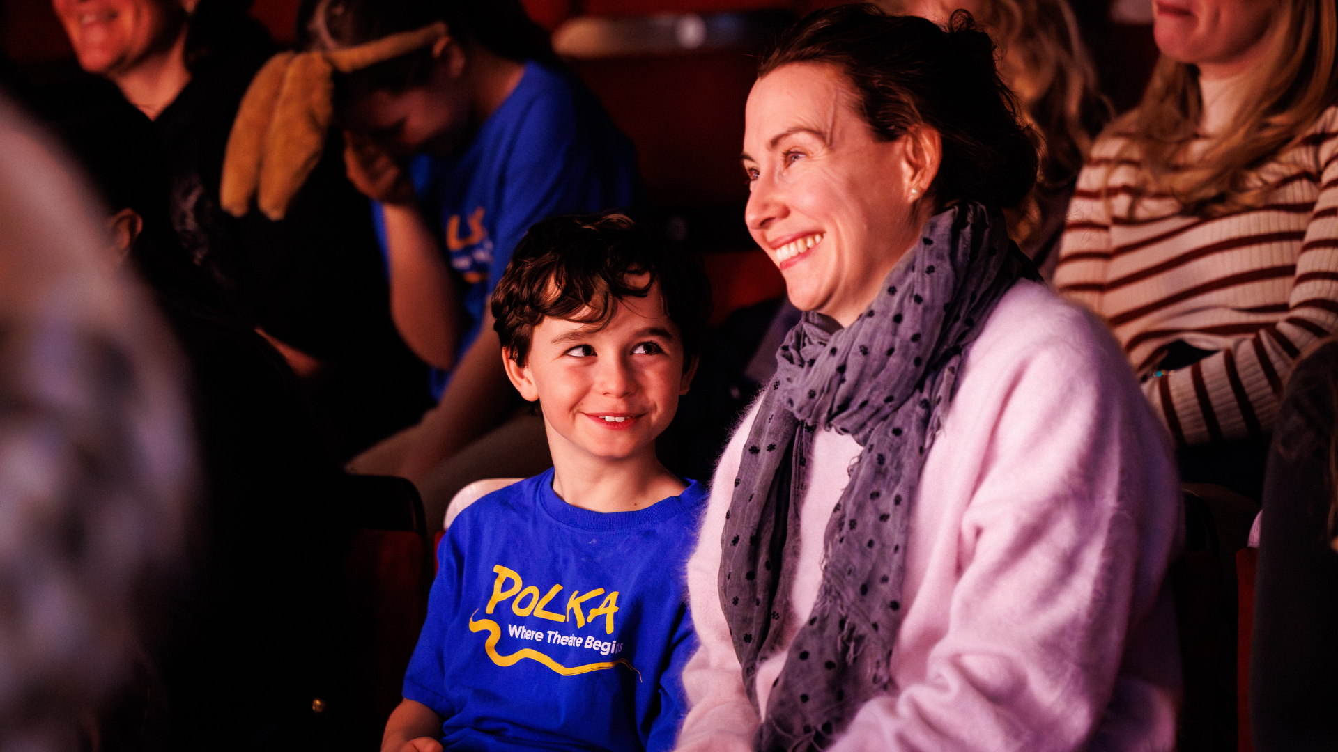 A mother and son sat in the audience at a performance, smiling happily. The son is wearing a blue Polka theatre t-shirt.