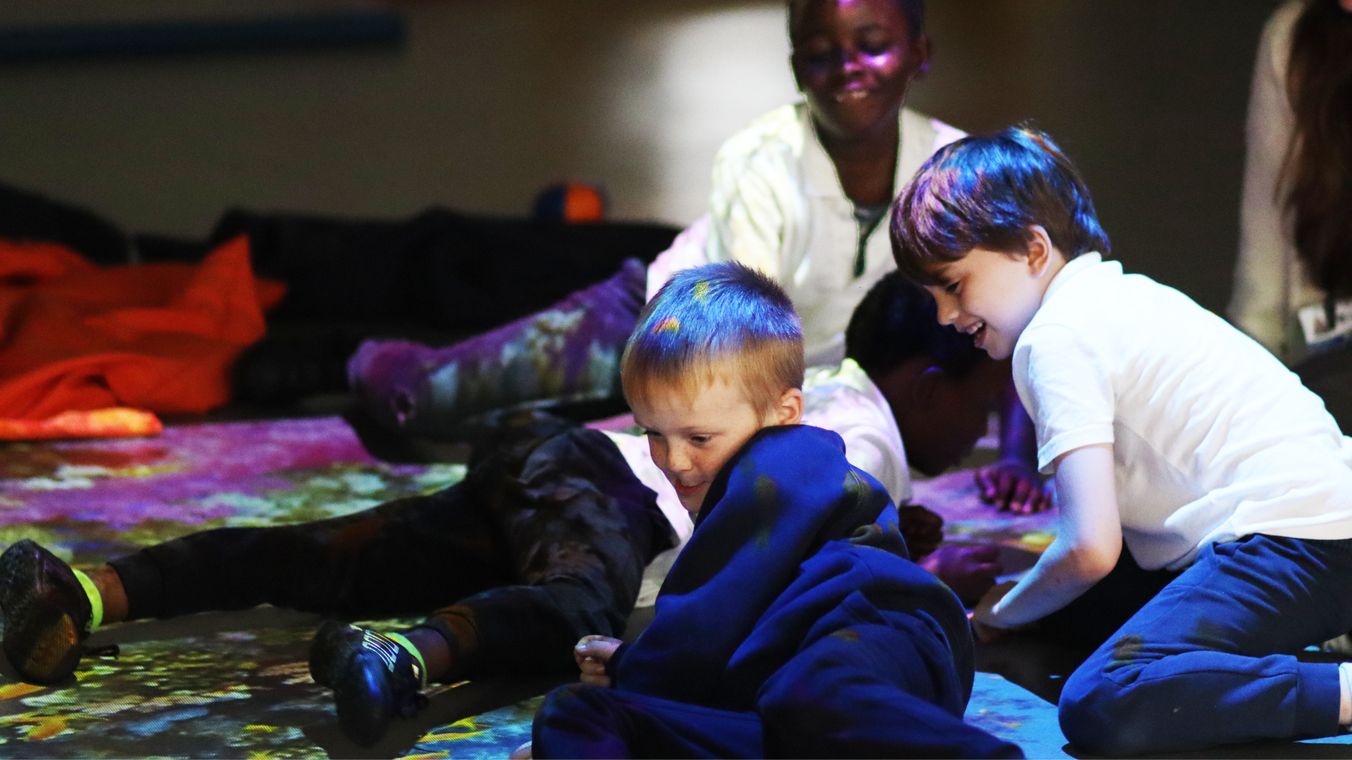 A group of primary school children lying on the floor during an activity - there is lighting projected over them.
