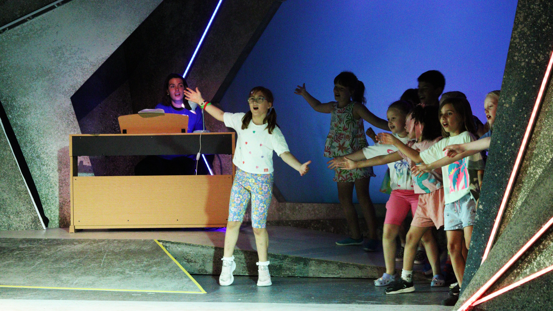 A group of children singing and dancing with a pianist on a wooden piano behind them.