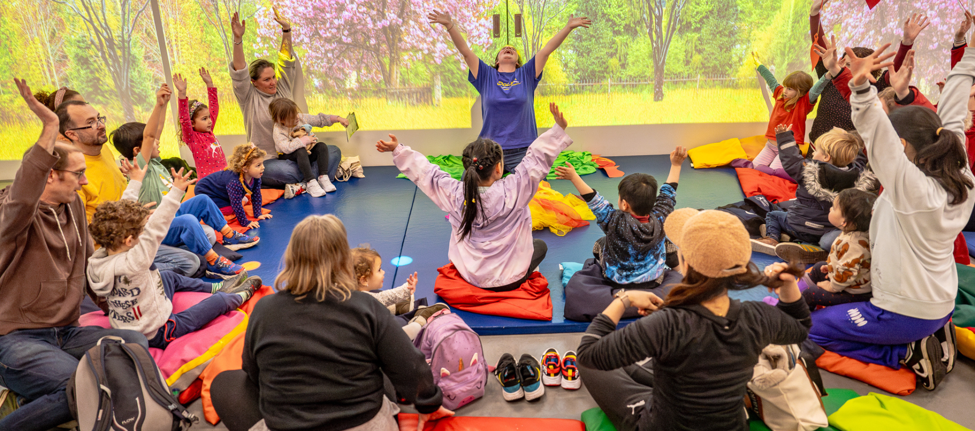 A Polka staff member holding a Saturday club sat on blue mats with a group of children. They are all holding their arms out upwards with their parents sat behind them.