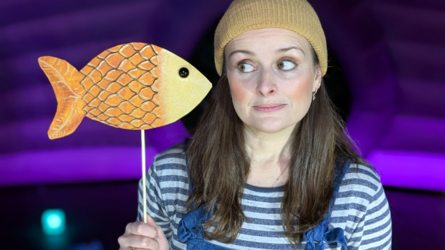 A performer wearing a striped t-shirt, yellow beanie and blue dungarees holding a paper fish on a stick.