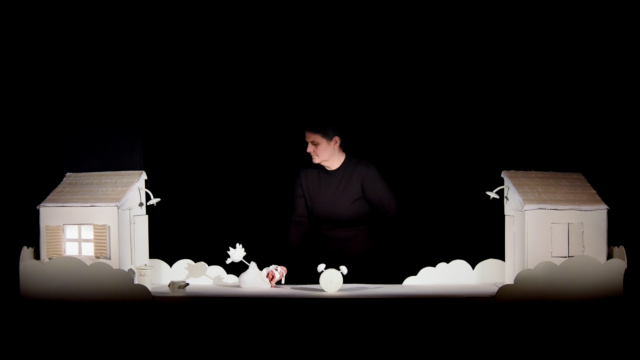 Two prop white houses with a white alarm clock in the middle of them, with a puppeteer holding a small bird on a stick.