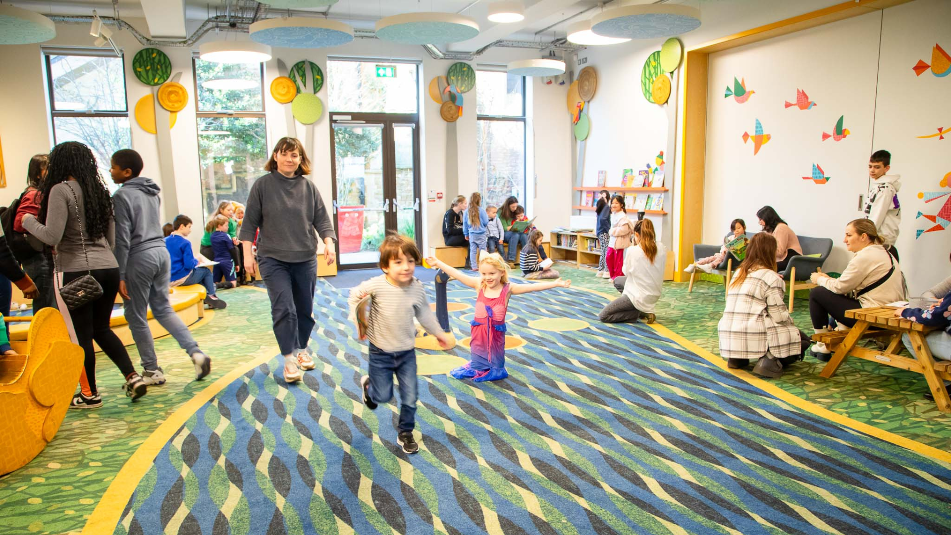 A group of children playing indoors in the play area under adult supervision.